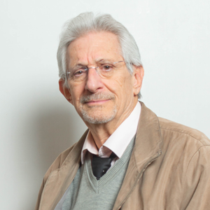 A man with short white hair and glasses wearing a brown blazer smiles towards the camera