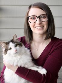 A woman with medium length straight brown hair and glasses smiles while holding a white and grey cat