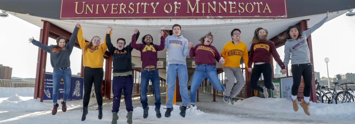 Group of people wearing University of Minnesota sweatshirts and jumping in front of a U of M sign
