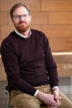 A man with short ginger hair and glasses sites and smiles towards the camera