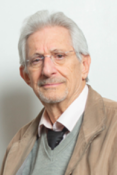 A man with short white hair and glasses wearing a brown blazer smiles towards the camera