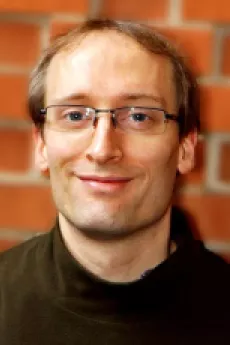 A man with short brown hair and glasseswearing a black turtleneck smiles towards the camera