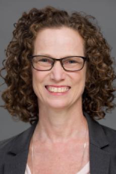 A woman with curly brown hair and glasses wearing a grey blazer smiles towards the camera