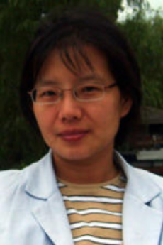 A woman with short black hair and wire framed glasses wearing a while lab coat and yellow and white striped shirt smiles towards the camera