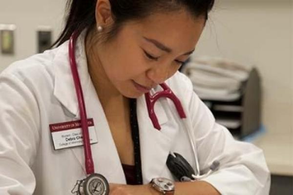 A VCS student wearing a white coat and red stethoscope looks at her watch