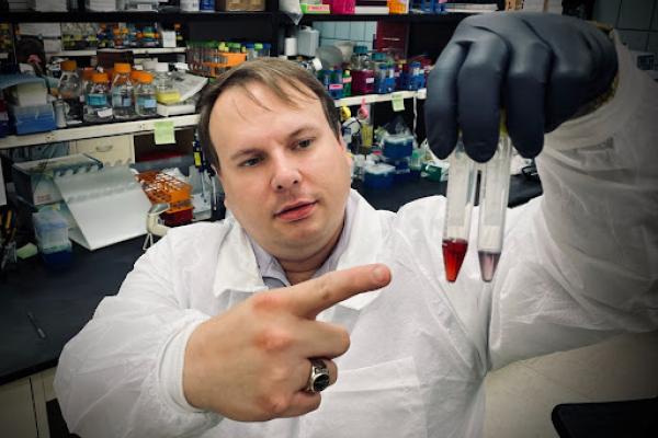 A man wearing a white lab coat and a black glove holds up two tubes and points to them