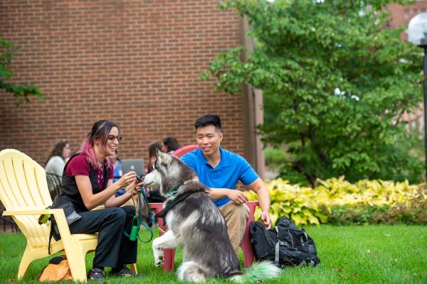 Two students sitting in chairs with a dog on the grass between them